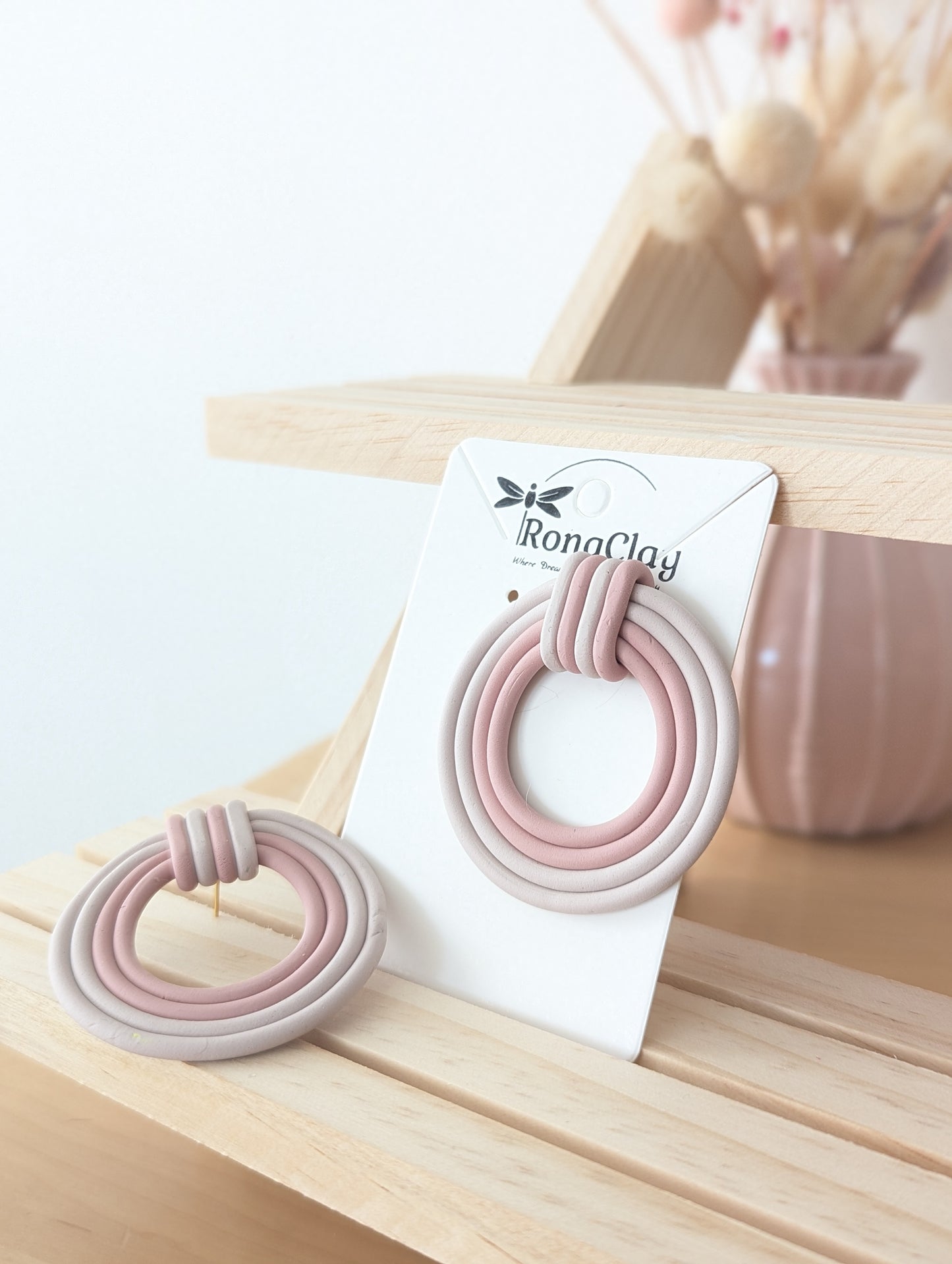 Handmade polymer clay earrings in pink and neutral tones, displayed on RonaClay branded packaging with a wooden background.